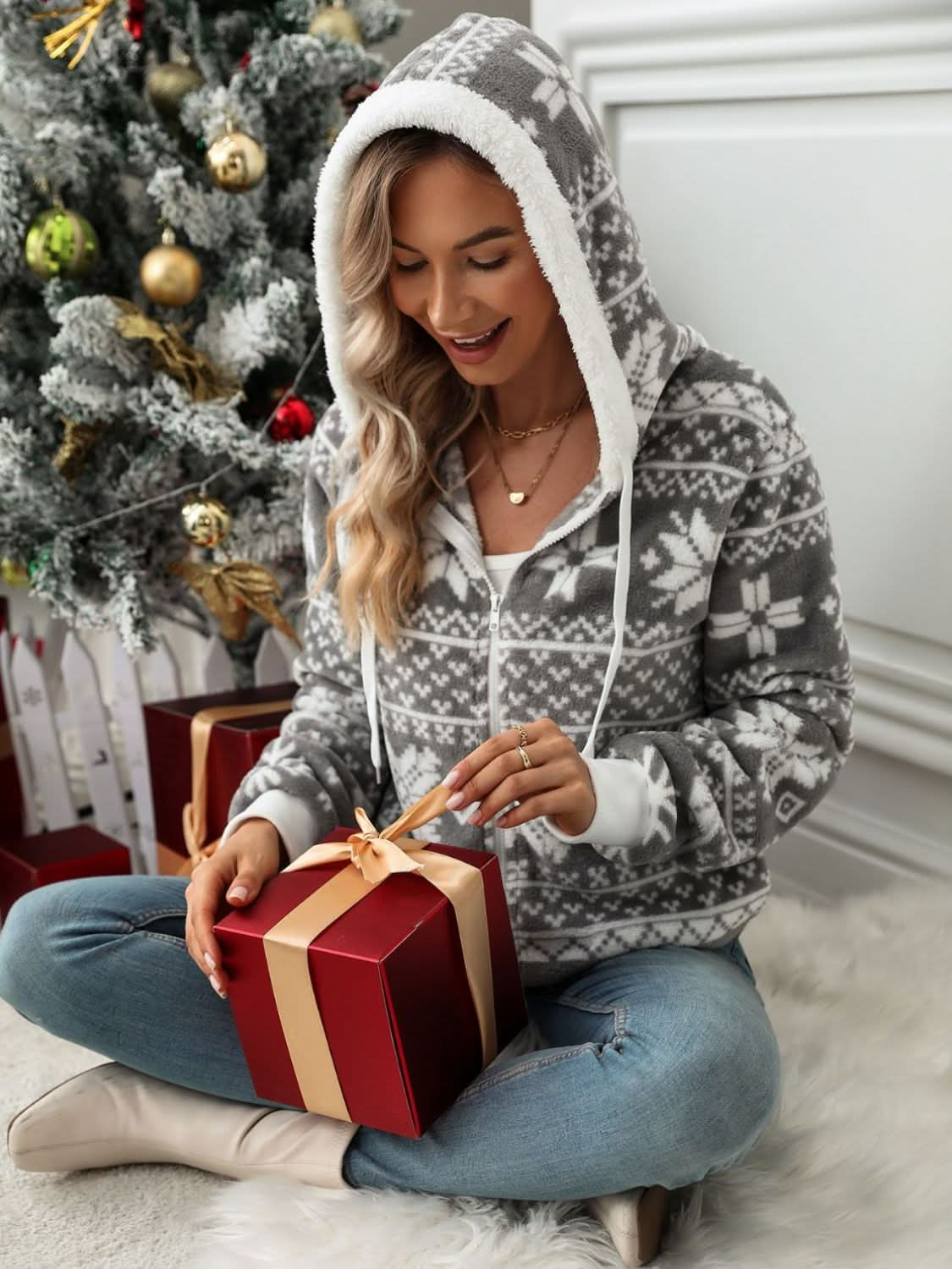 Drawstring snowflake zip up hooded outerwear worn by woman sitting near Christmas tree, holding a wrapped gift.