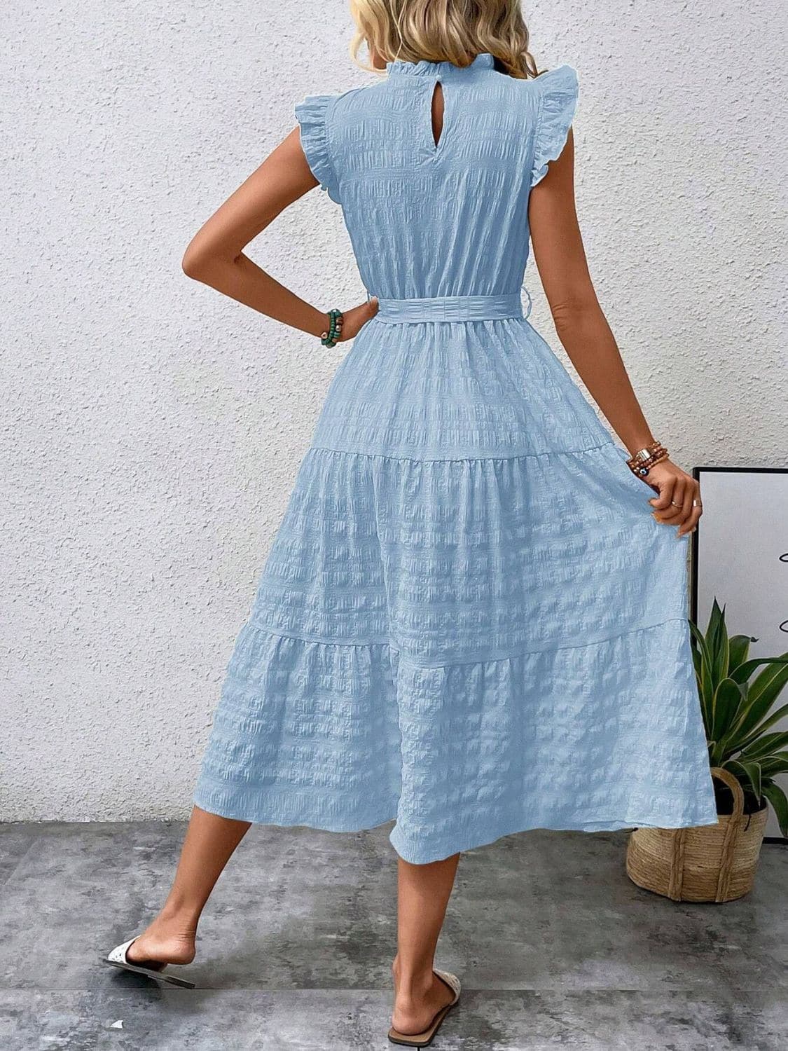 Ruffled cap sleeve midi dress in light blue with tie detail, worn by a woman standing by a plant.