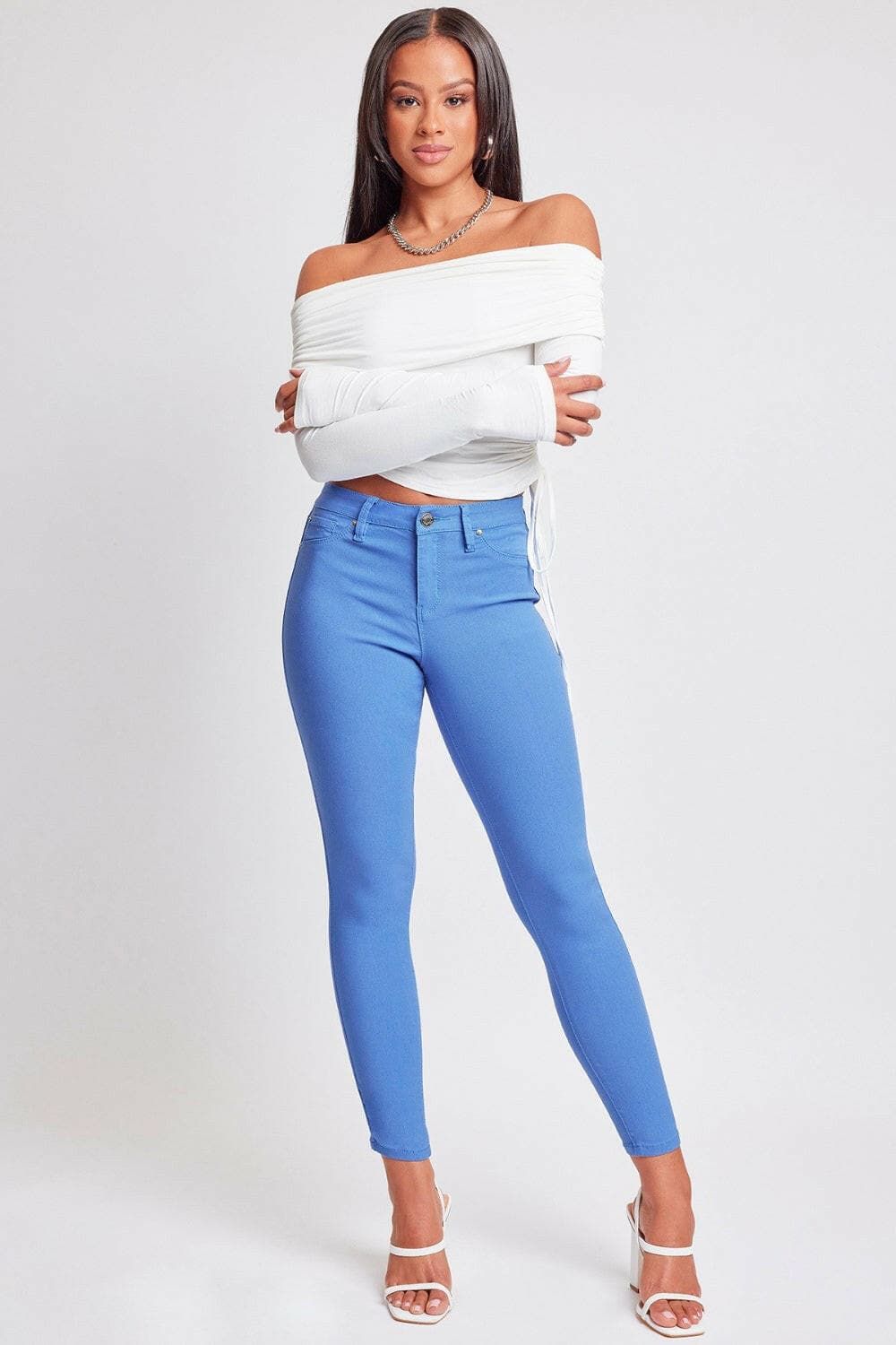 YMI Jeanswear Full Size Hyperstretch Mid-Rise Skinny Pants in blue, styled with a white off-shoulder top and white heels.