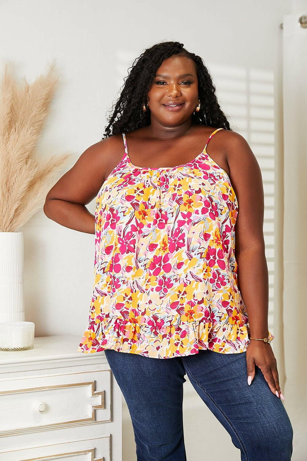 Plus-size model wearing a sleeveless floral ruffle hem top, featuring a vibrant floral design and scoop neck, paired with jeans.